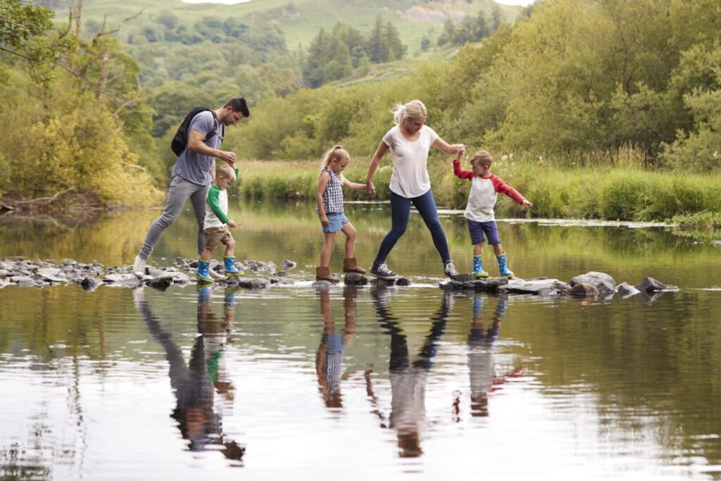 Family Crossing River Whilst Hiking In UK Lake District