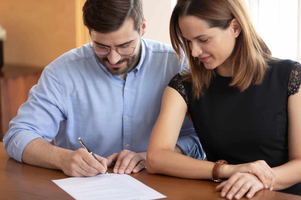 Pleasant young woman watching smiling husband signing contract.