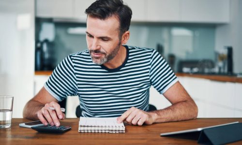 Smiling man managing home budget with calculator and notebook while sitting at dining table