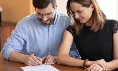Head shot close up pleasant young woman watching smiling husband signing contract. Happy family couple making first house purchase deal. Excited clients putting signature on banking agreement.
